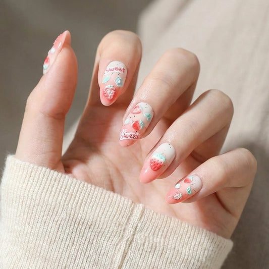 Stylish almond-shaped nails with a pink and white ombre design, featuring hand-painted strawberries, hearts, and "sweet" lettering, modeled against an off-white knit sweater background.