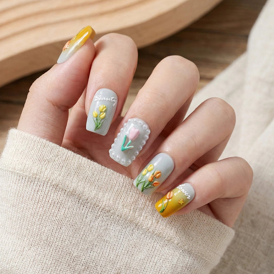 Stylish 3D floral nail art with a light gray base, hand-painted tulips, pearl detailing, and yellow gradients, presented against an off-white knit sweater background.