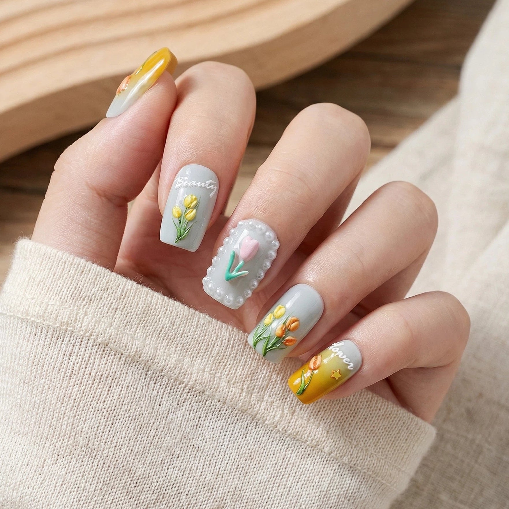 Stylish 3D floral nail art with a light gray base, hand-painted tulips, pearl detailing, and yellow gradients, presented against an off-white knit sweater background.